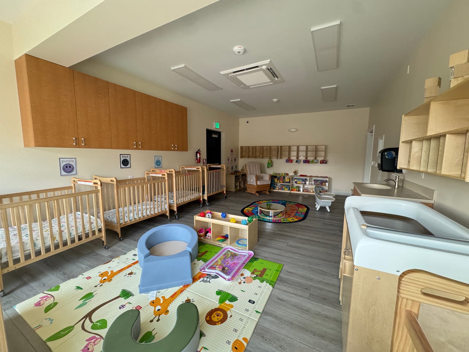 Infant room at the Midway Blvd location with a row of cribs, a glider, a padded play mat, and a changing station
