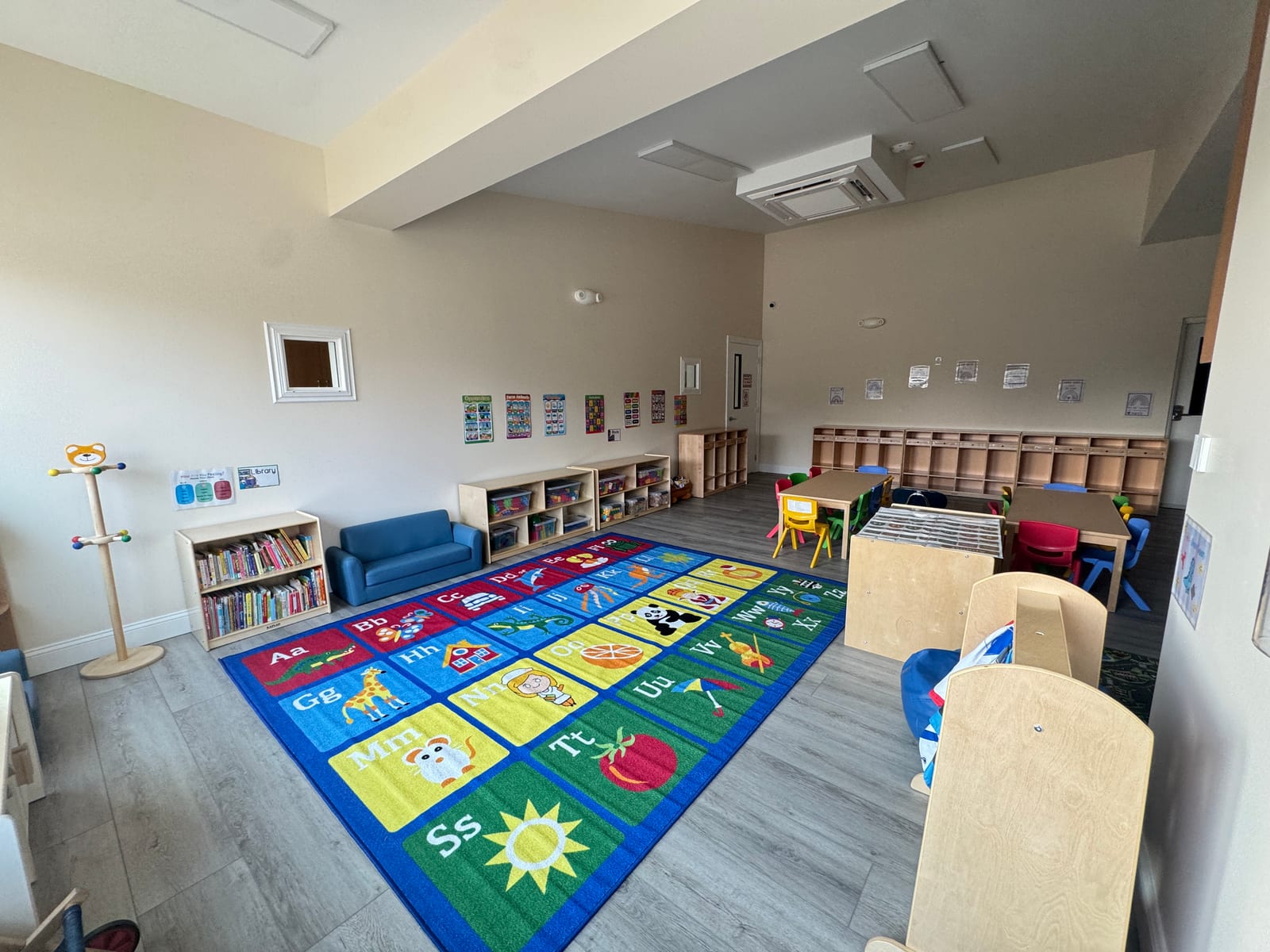 Toddler classroom at the Midway Blvd location with an alphabet rug, a library corner, a reading couch, activity tables, and a wall of cubbies
